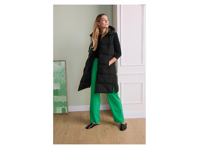 Vrouw in lange zwarte bodywarmer met capuchon, zwarte blouse en groene broek.