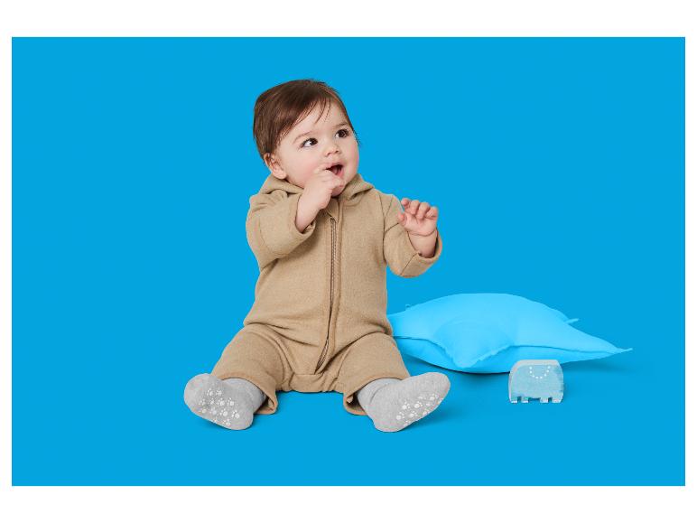 Schattige peuter in een beige jumpsuit zit naast een blauw kussen en een houten speeltje.