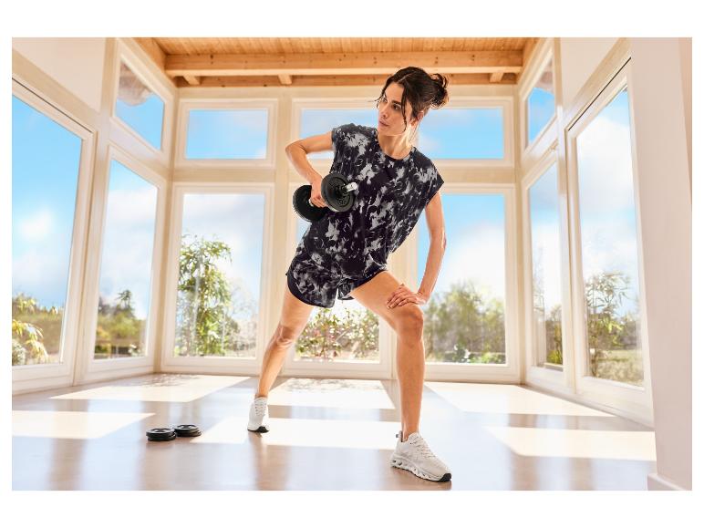 Vrouw in CRIVIT sportshort traint met een dumbbell in een lichte kamer.