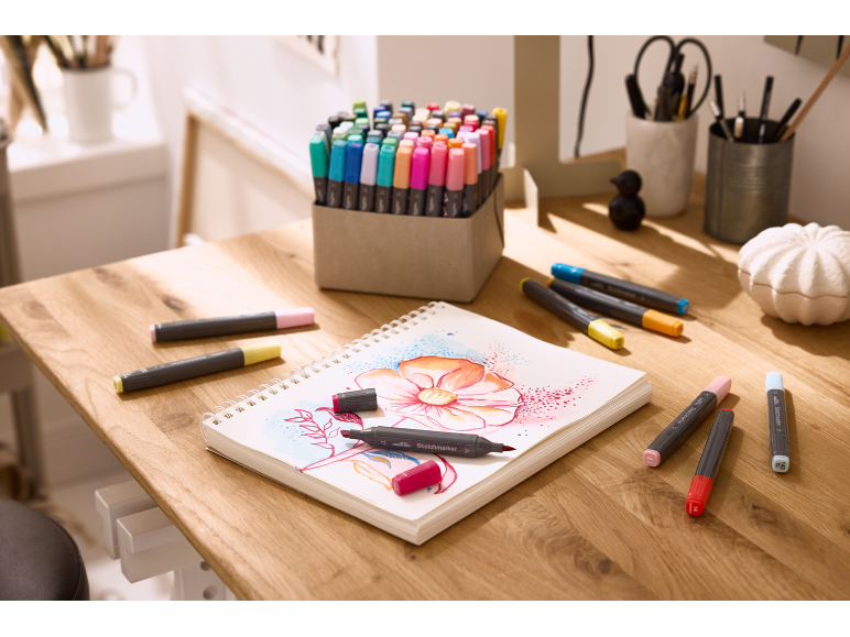 Een houten tafel met een schetsboek met bloemtekening en diverse stiften.