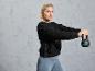 Een vrouw in een zwarte trui en grijze leggings houdt een zwarte kettlebell vast.
