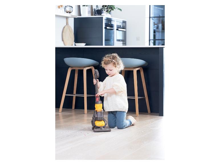 Een klein meisje stoft de vloer met een speelgoed Dyson stofzuiger.