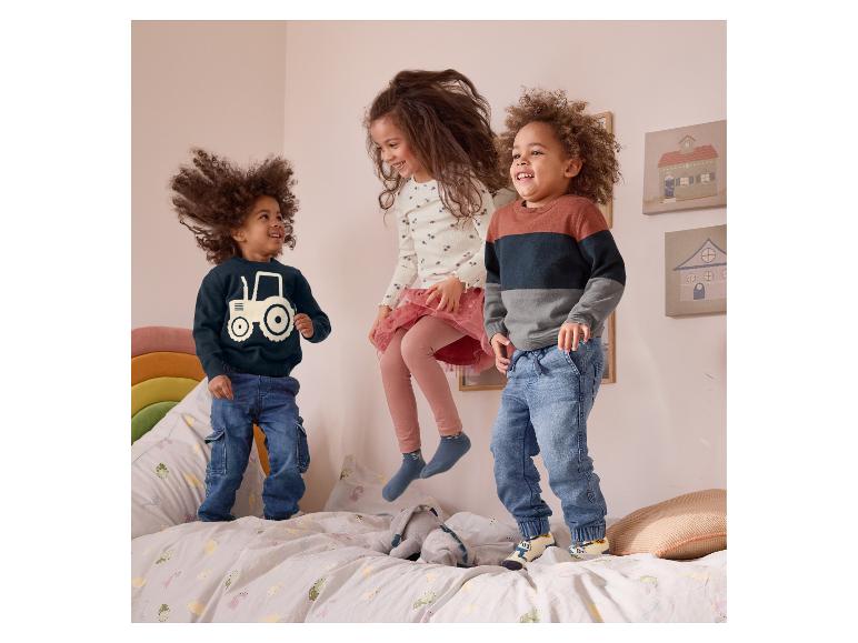 Drie kinderen springen op een bed met kinderbeddengoed, gekleed in leuke truien en sokken.