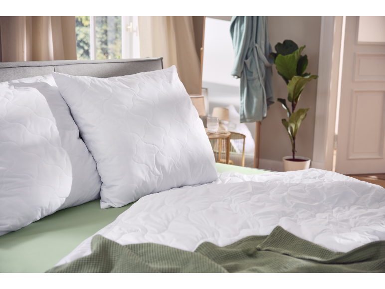 Witte doorgestikte kussens en dekbed op een bed met groene lakens en deken.