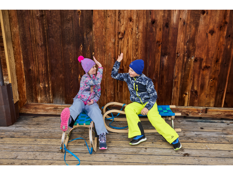 Twee kinderen in winterkleding en mutsen, zittend op sleeën en high-fivend.