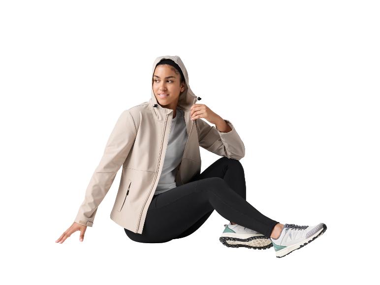 Vrouw in beige jas met capuchon, zwarte legging en sneakers, zittend.