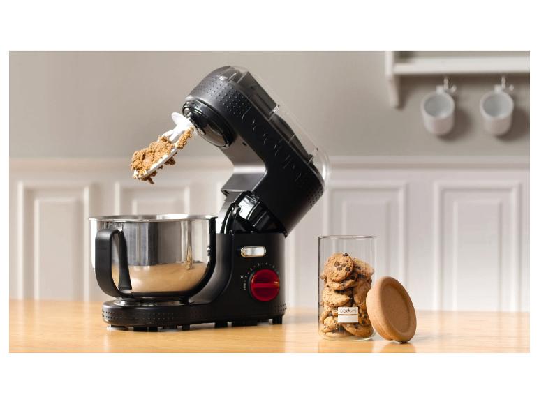 Een zwarte keukenmachine met deeg in de kom en een pot koekjes op een houten tafel.