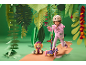 Meisje met helm op een step, naast een knuffelhond, in een jungle-achtige setting.