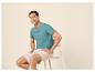 Man in turquoise T-shirt en beige korte broek zit op een kruk.