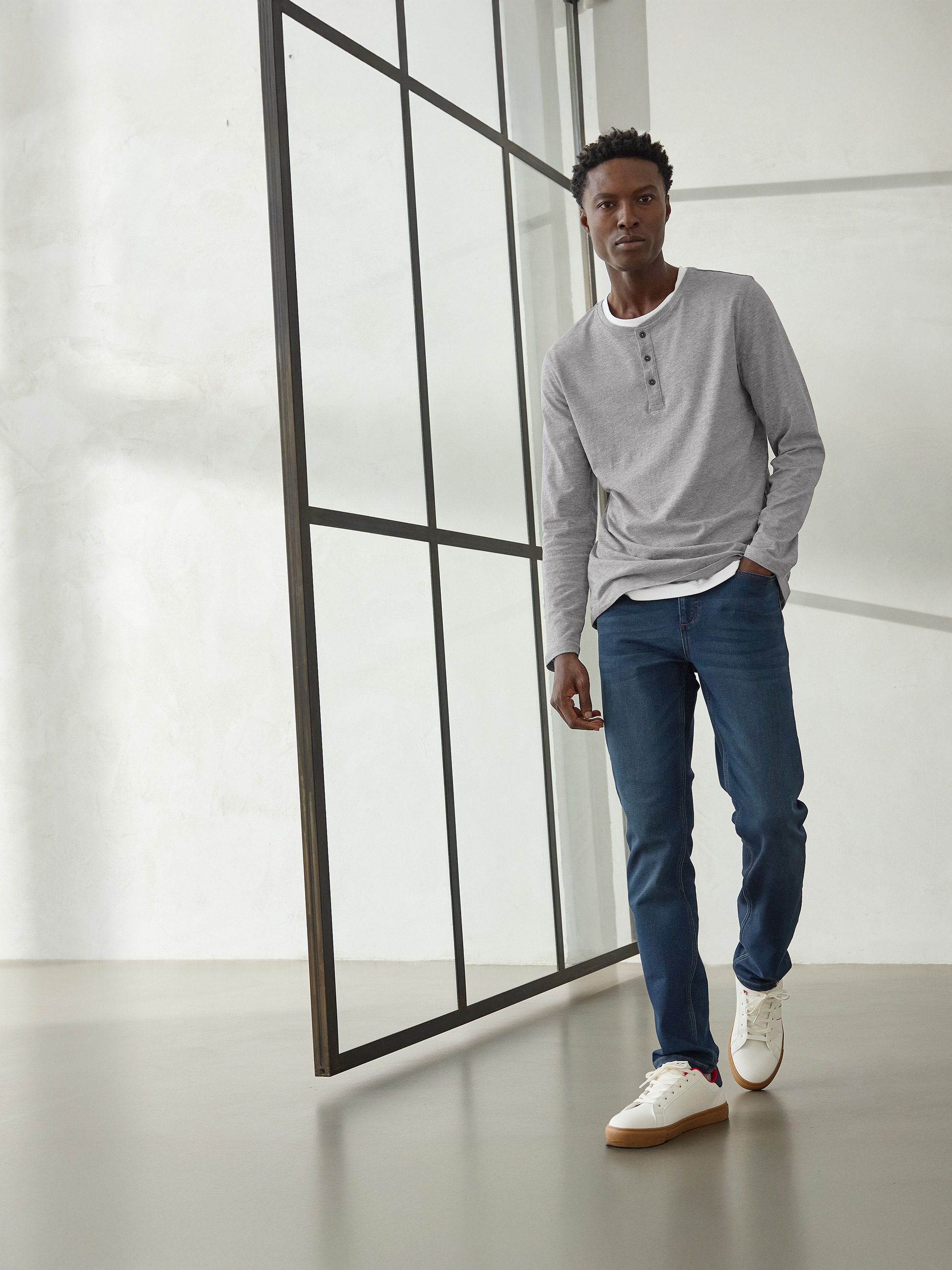 Man in grijs Henley-shirt met lange mouwen, donkerblauwe jeans en witte sneakers.
