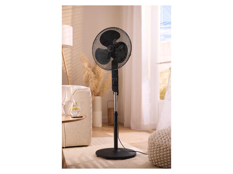 Zwarte staande ventilator in een woonkamer met een bank, lamp en raam.