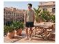 Man in beige poloshirt en donkere shorts op een zonnig terras met uitzicht op de stad.