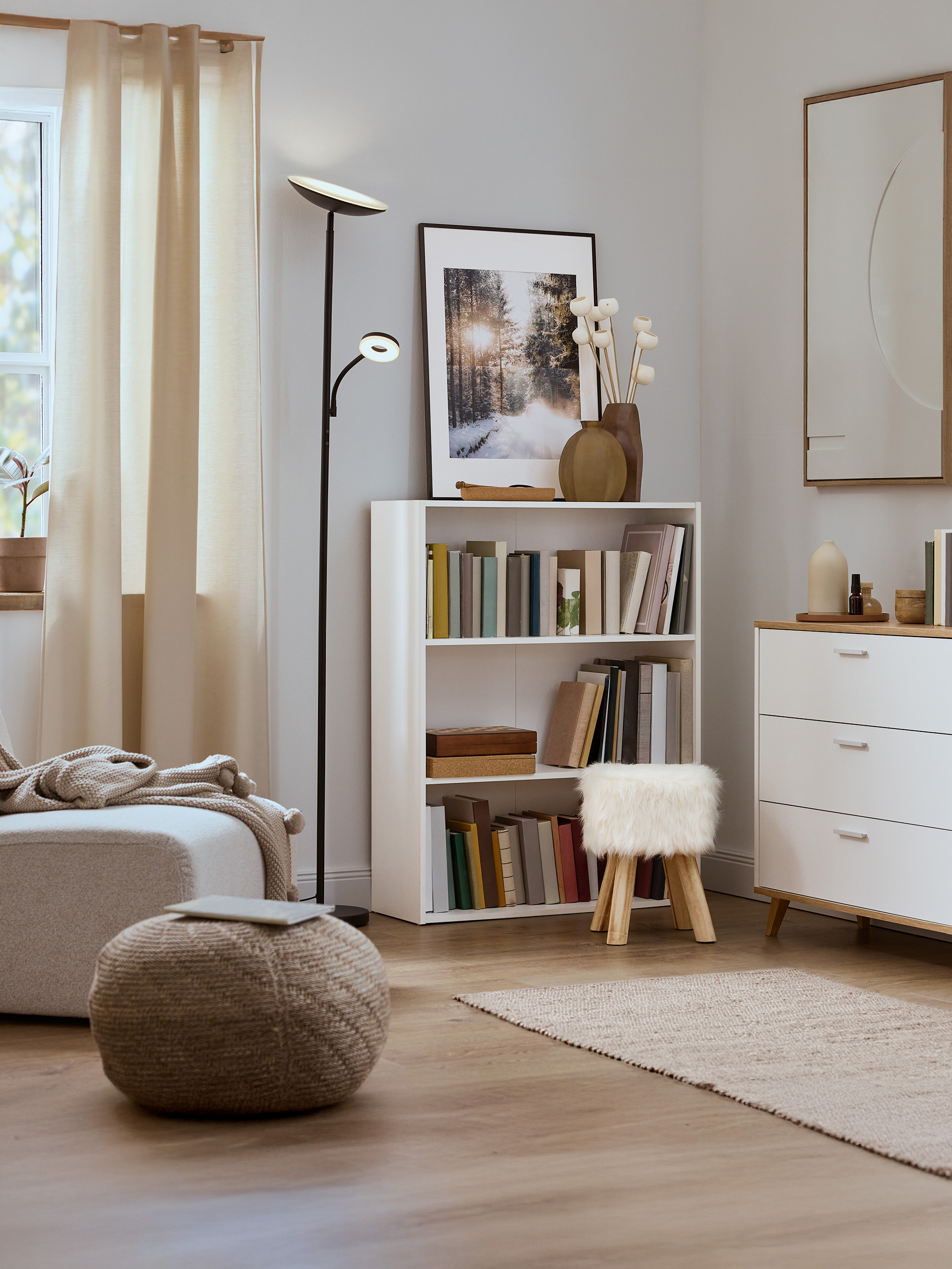 Lichte woonkamer met witte boekenkast, gebreide poef en moderne staande lamp.