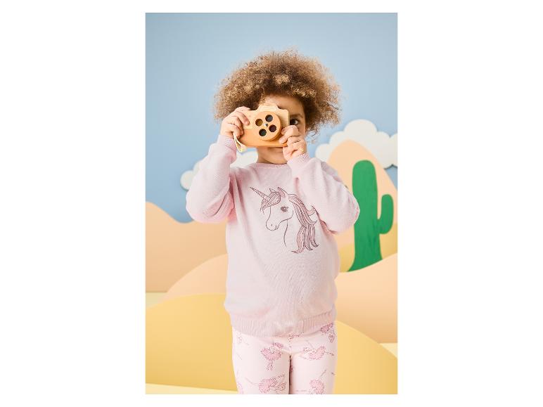 Meisje met krullend haar in roze trui met eenhoorn en roze broek met ballerina's, speelt met houten camera.
