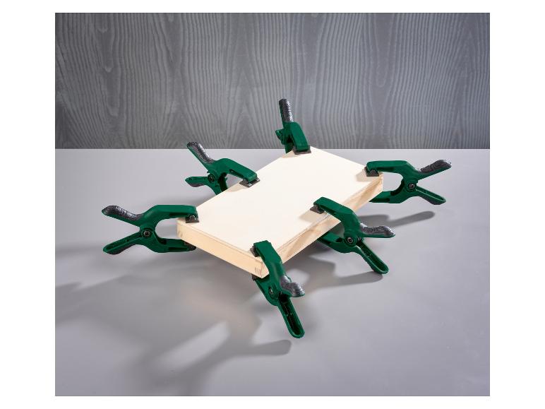 Zes groene veerklemmen houden een houten frame vast op een grijze achtergrond.