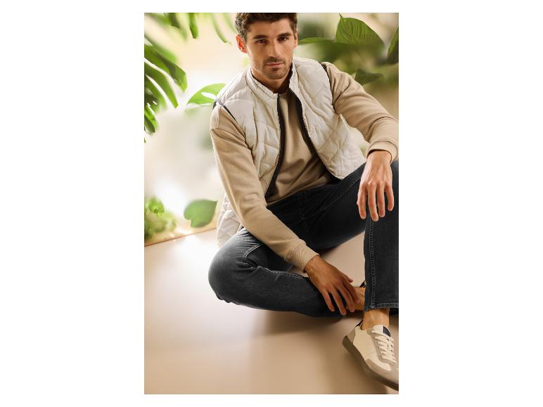 Man in beige gewatteerd vest, sweatshirt en donkere jeans, zittend in een natuurlijke omgeving.