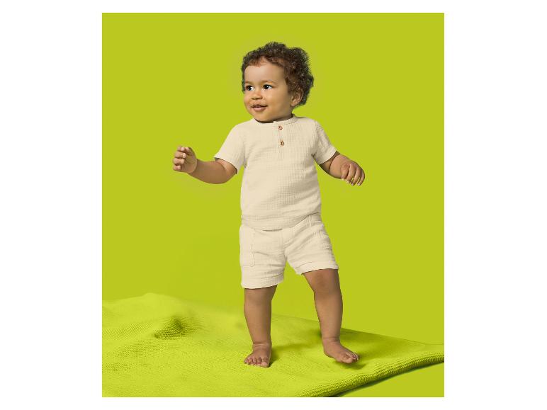 Peuter in een crèmekleurig shirt met korte mouwen en korte broek, staand op een groene mat.