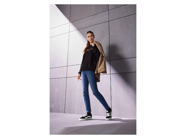 Vrouw in zwarte trui met lange mouwen, jeans en zwarte sneakers, met beige jas over haar schouder.