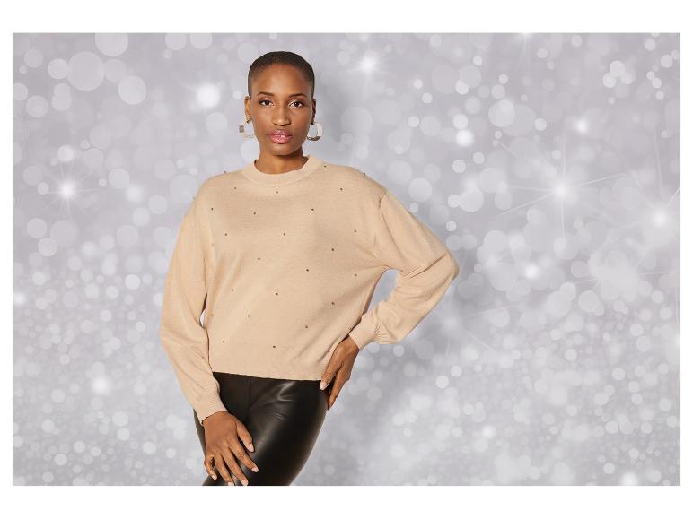 Een vrouw draagt een beige trui met glinsterende details en een zwarte leren broek.