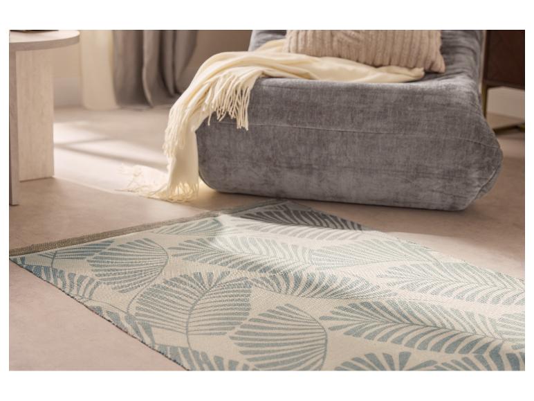 Grijze fauteuil met beige plaid en kussen, licht vloerkleed met blauw bladpatroon op de vloer.