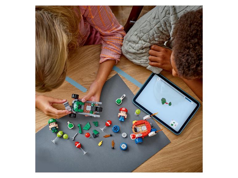 Twee kinderen spelen met LEGO Super Mario sets, waaronder een kart en figuren, begeleid door een tablet.