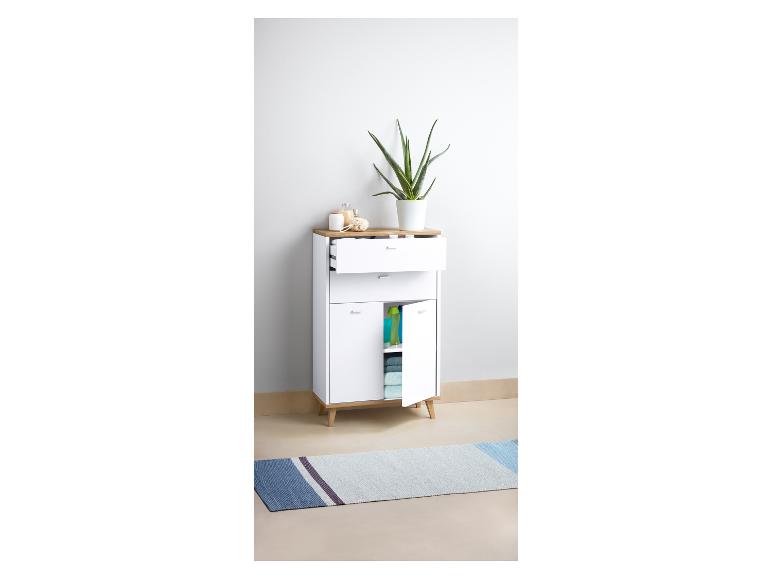 Een wit badkamerkastje met een lade en twee deuren, een plant, een badmat en badaccessoires