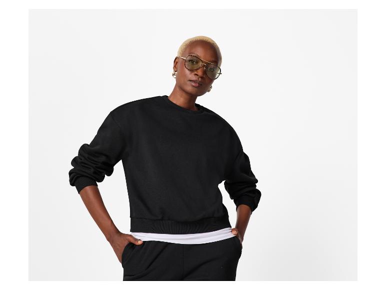 Vrouw in zwart sweatshirt en joggingbroek, met handen in zakken.
