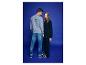 Man in grijze sweater en jeans, vrouw in zwarte sweater en broek tegen blauwe achtergrond.