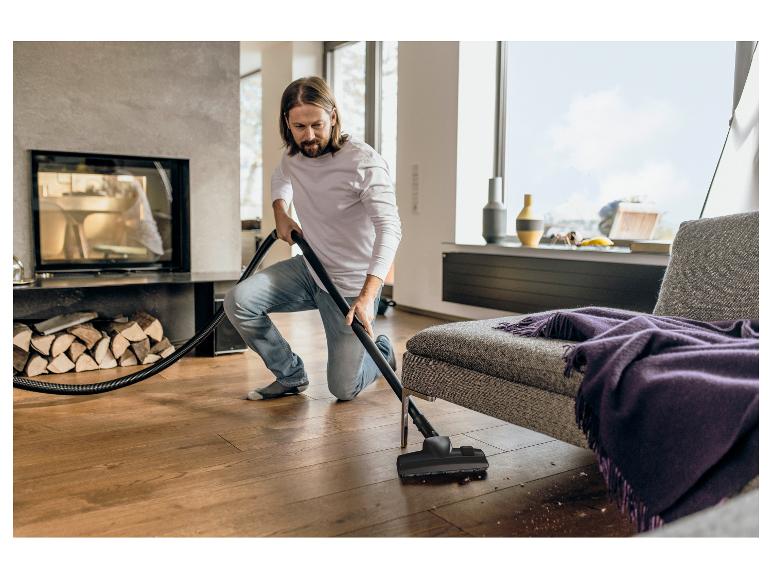 Man stofzuigt houten vloer, met open haard en bank op de achtergrond.