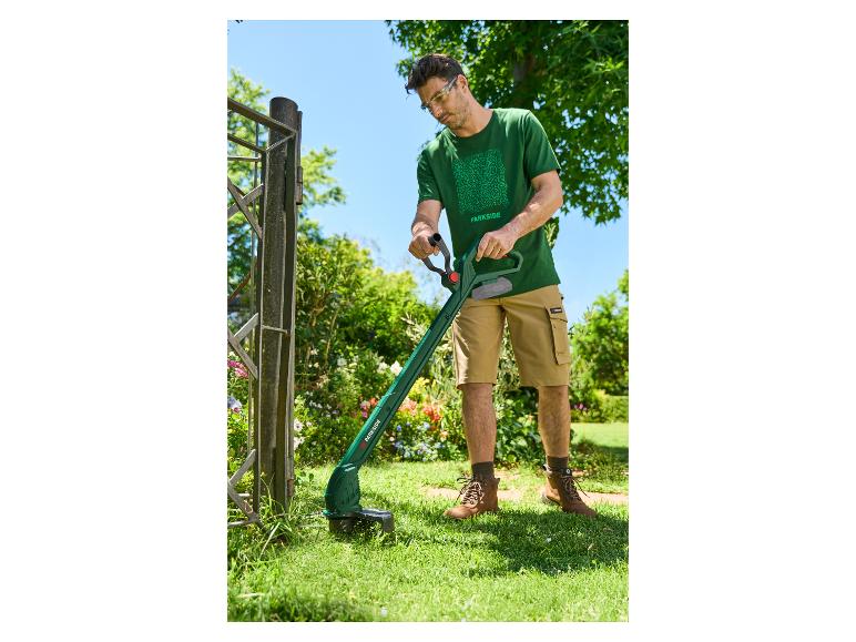 Man in groen T-shirt en korte broek gebruikt grastrimmer bij een hek.