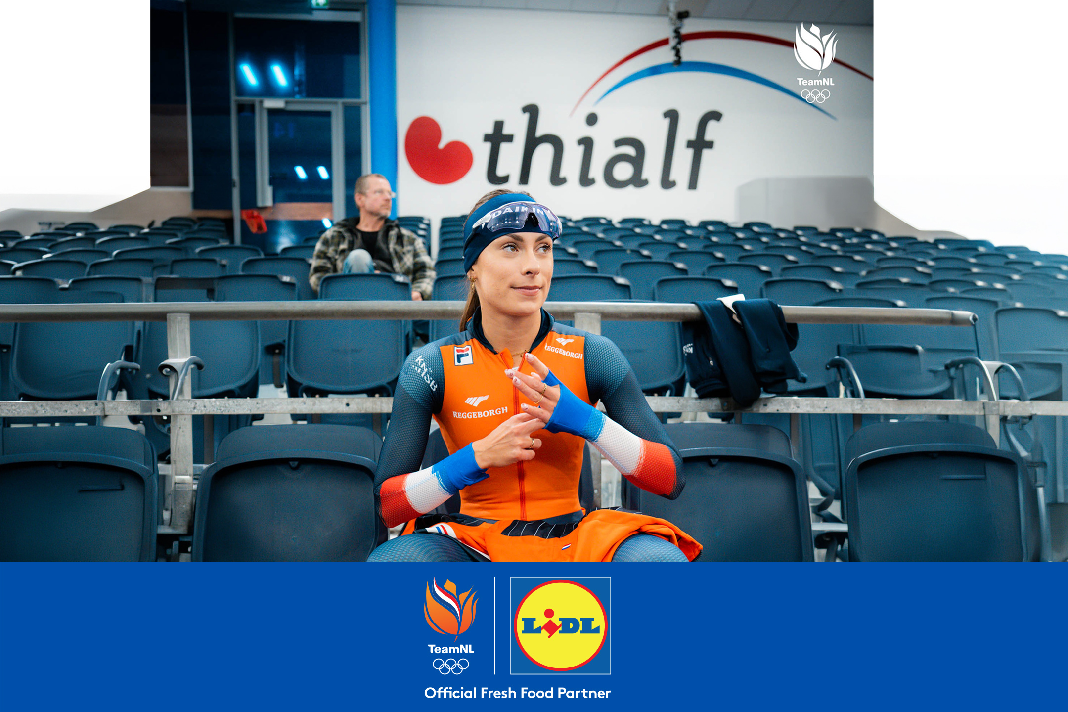 Een atlete in een oranje schaatspak zit op een tribune, met logo's van een sportteam en een supermarkt.