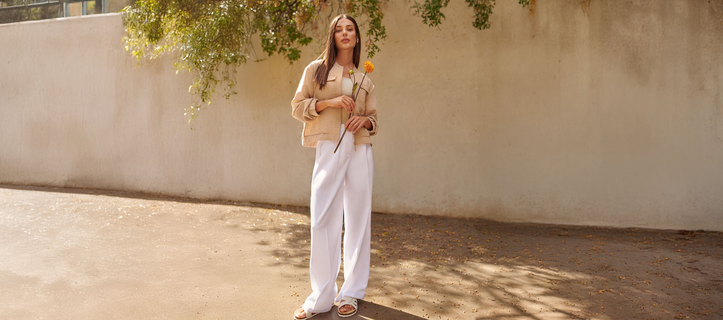Vrouw in beige jasje, witte broek en sandalen, met een gele bloem.
