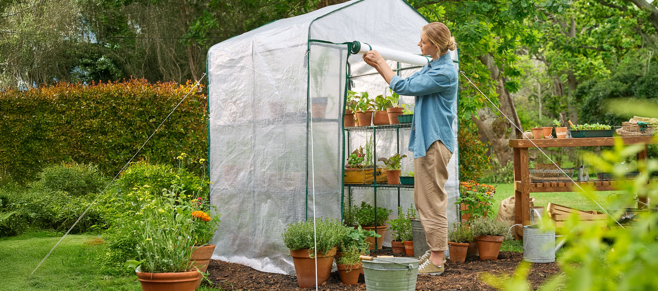 Vrouw opent een kas met planten en potten in een tuin.