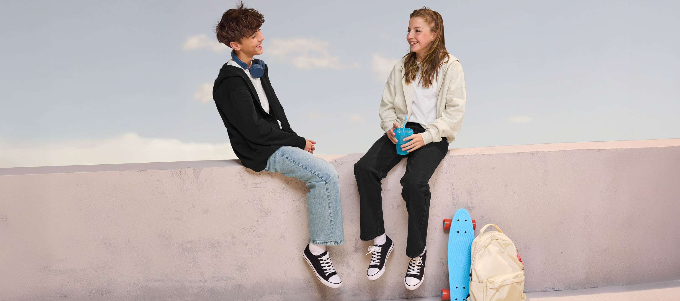 Twee tieners in hoodies en jeans zitten op een muur, met een skateboard en rugzak in de buurt.