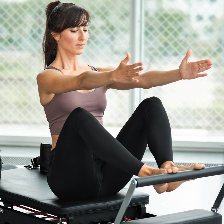 Vrouw doet Pilates op een reformer, draagt een top en zwarte legging.