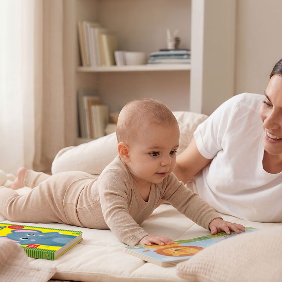 Baby in beige romper speelt met kleurrijke dierenboeken op een mat, met een lachende volwassene.