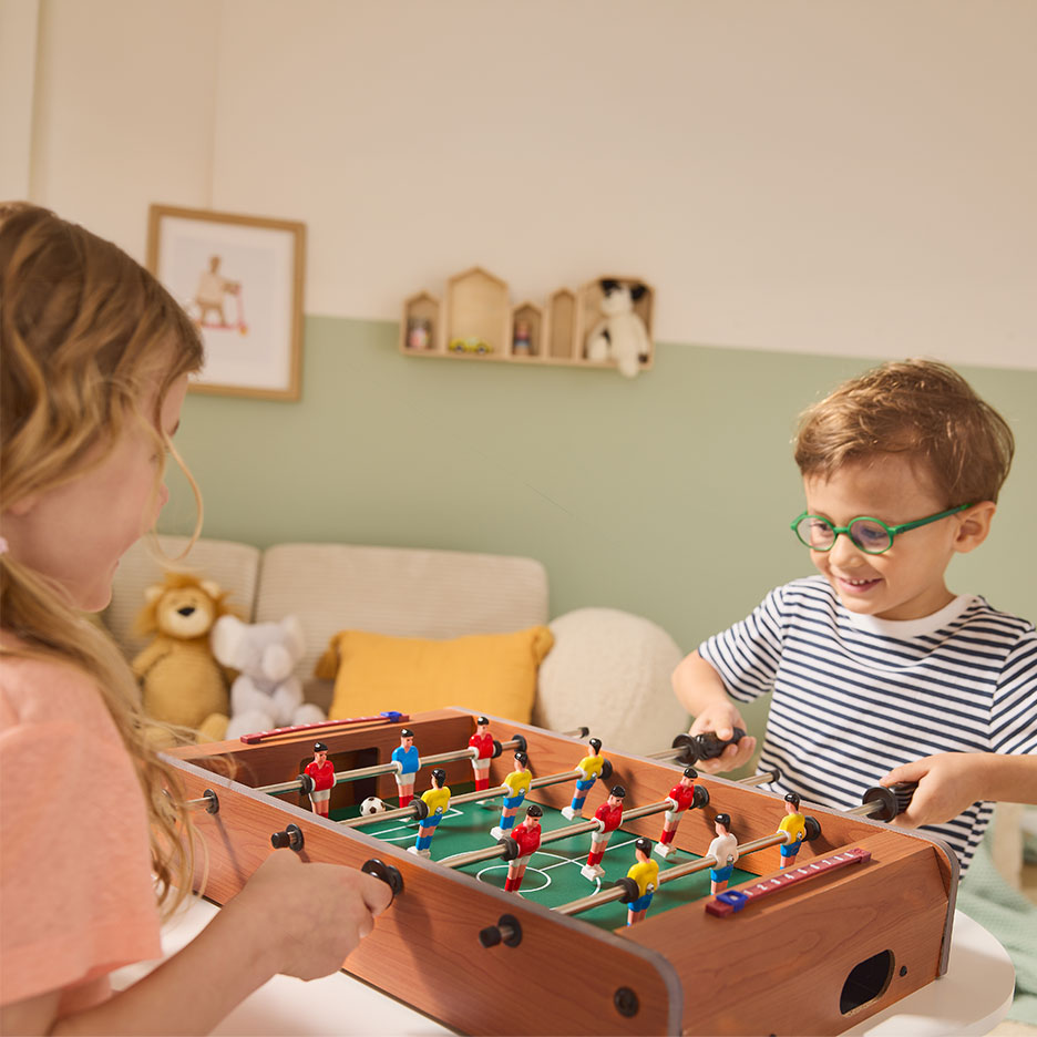 Twee kinderen spelen tafelvoetbal, meisje links, jongen rechts.