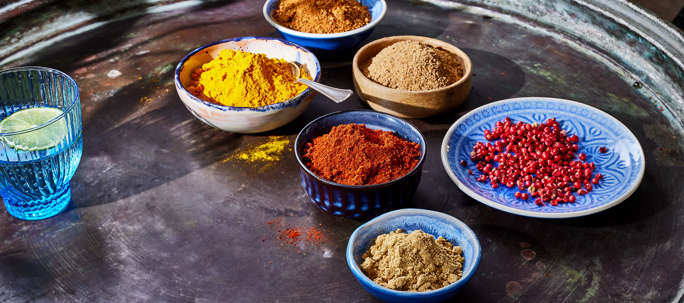 Diverse kruiden, zoals kurkuma, paprikapoeder en roze peper, in kommen op een donkere ondergrond.