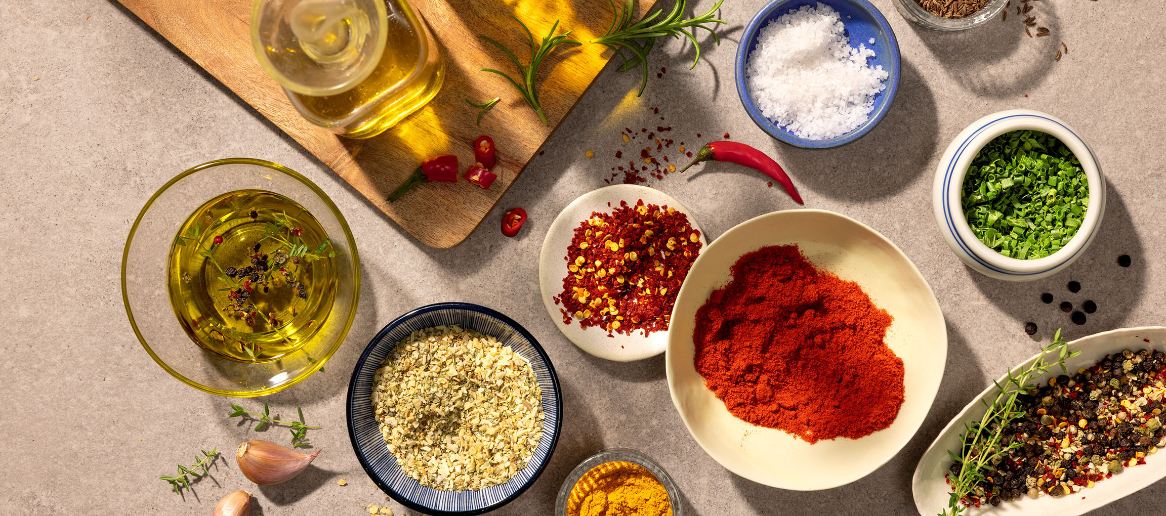 Diverse kruiden en oliën, waaronder paprika, chili, zout, peper en olijfolie, op een grijze achtergrond.