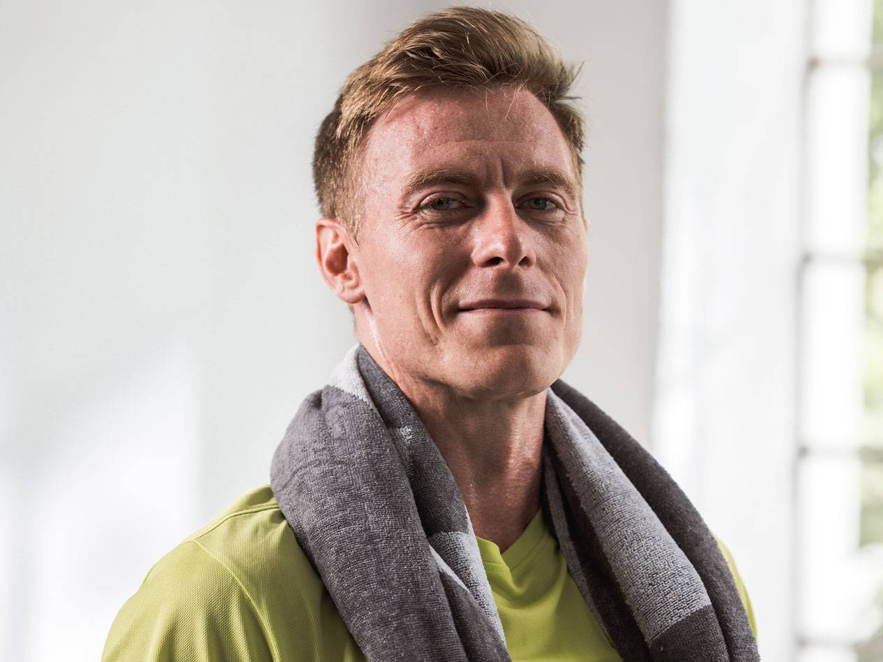 Een blonde man met een handdoek om zijn nek kijkt naar de camera.