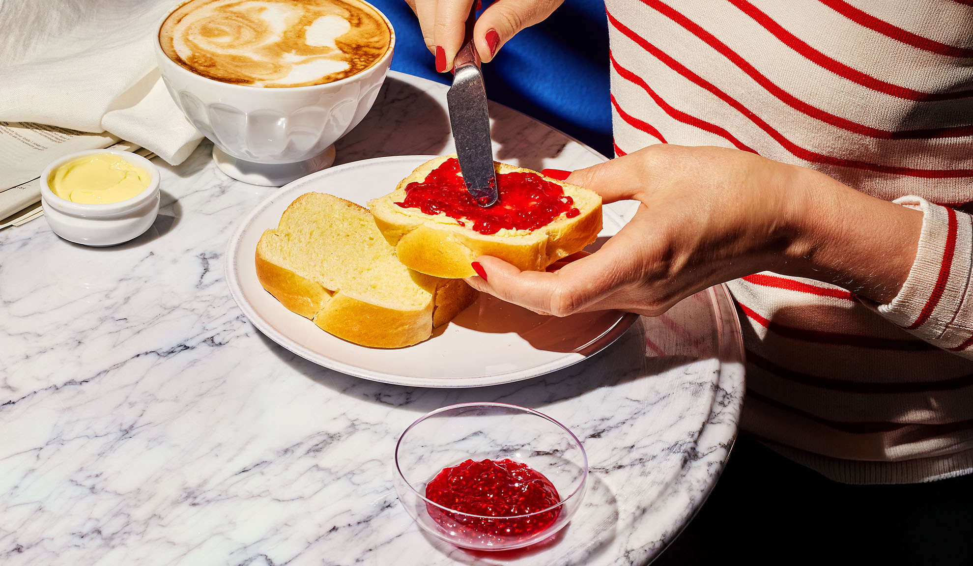 Handen smeren jam op briochebrood, met koffie en boter op een marmeren tafel.