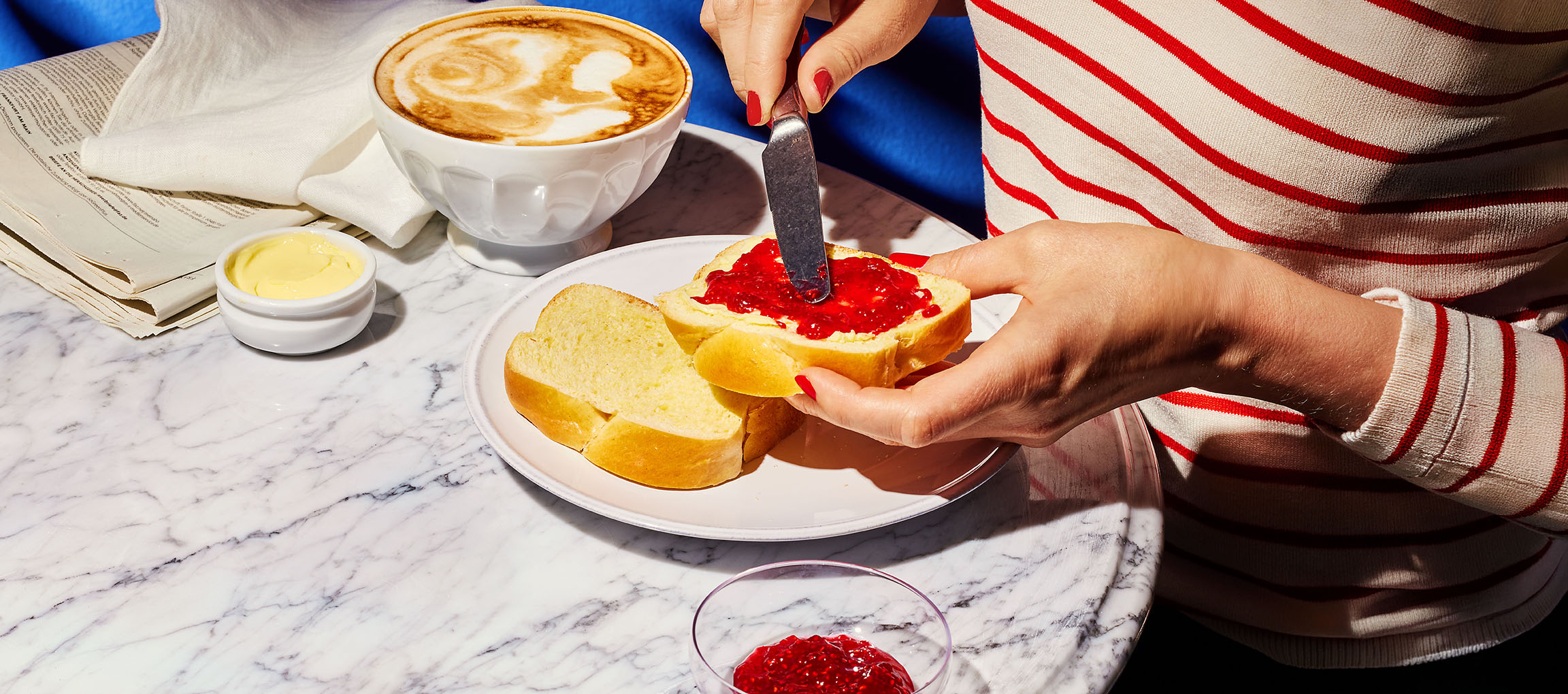 Handen smeren jam op briochebrood, met koffie en boter op een marmeren tafel.