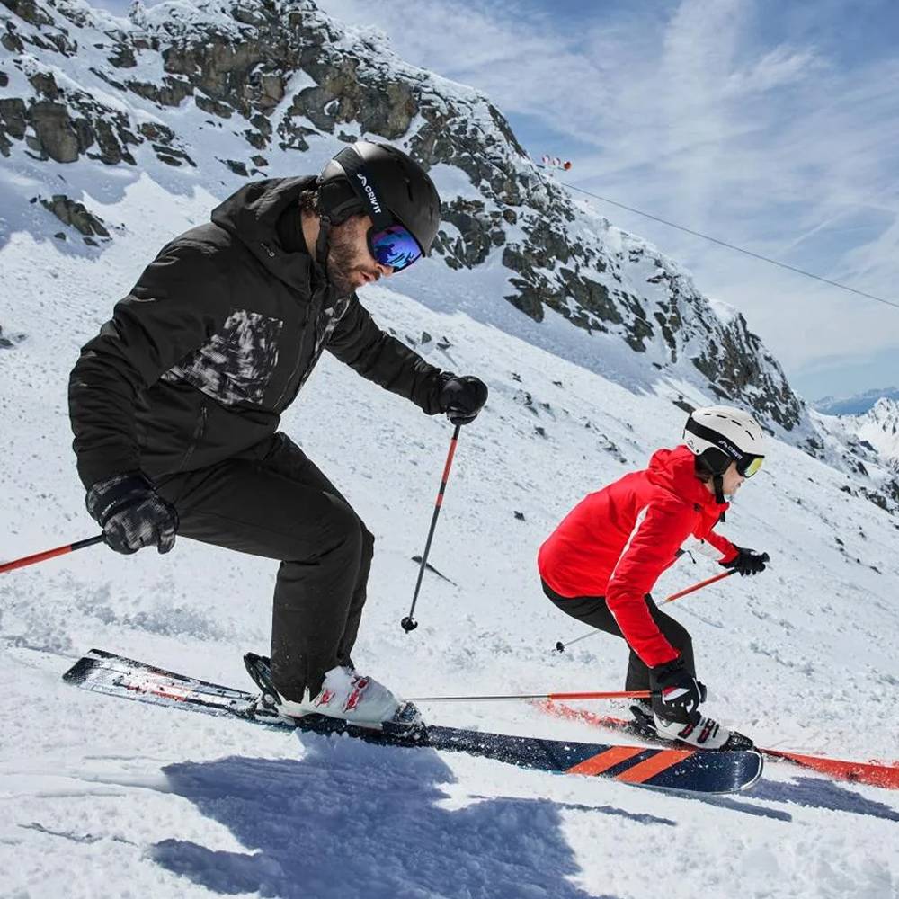 Twee skiërs in zwarte en rode ski-jassen, helmen en skibrillen op een besneeuwde berg.
