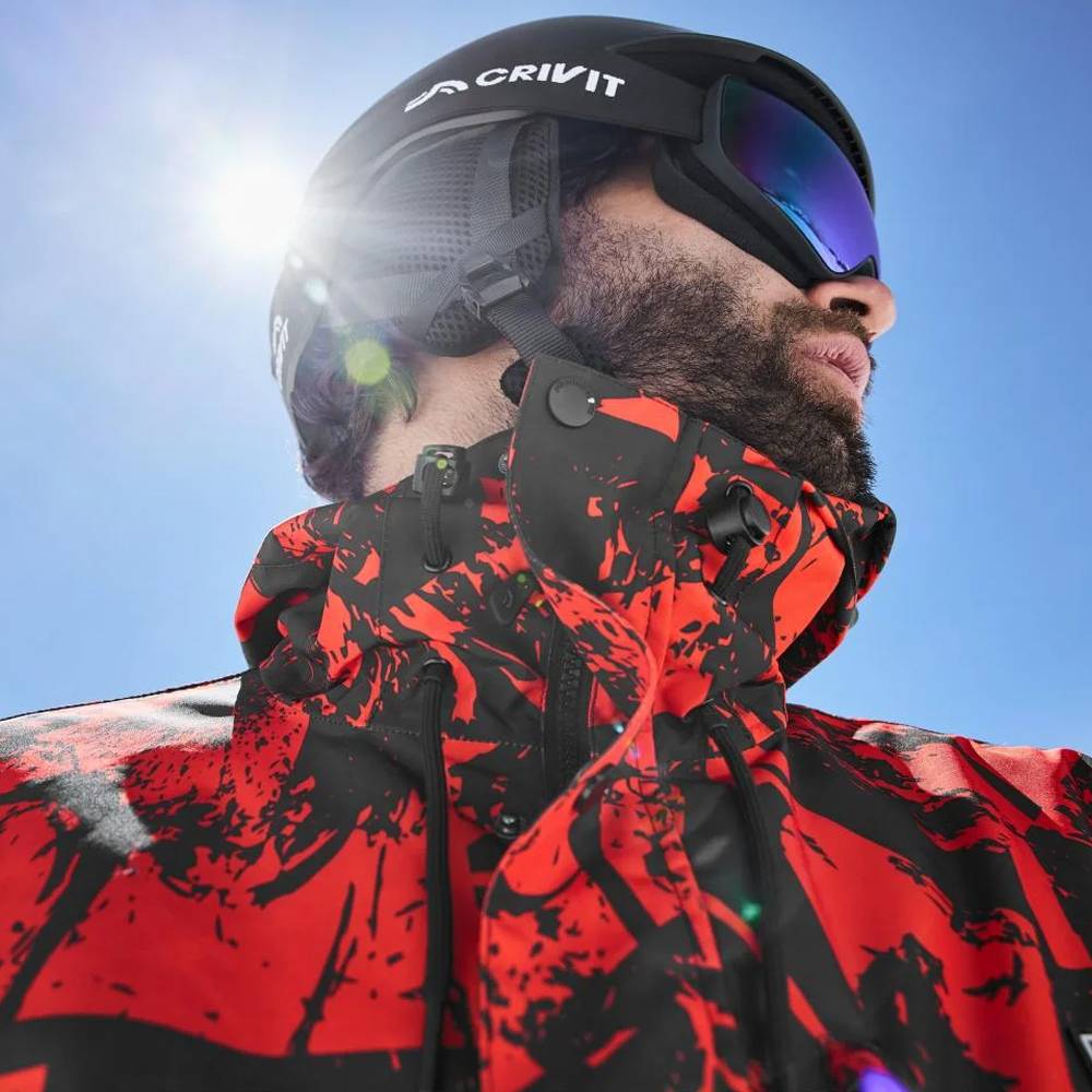 Man met Crivit skihelm, skibril en rood-zwart ski-jack onder een zonnige blauwe lucht.