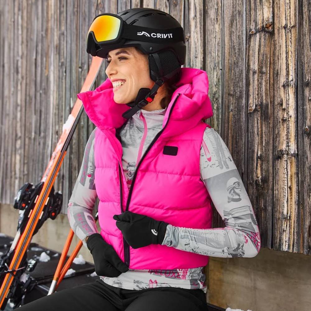 Vrouw in roze Crivit ski-vest, helm en skibril, lachend naast ski's.
