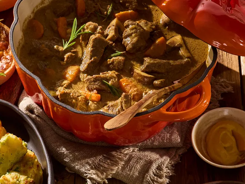 Stoofpot met vlees, wortels en rozemarijn in een oranje pan op een houten tafel.