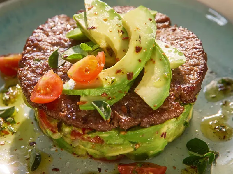Gestapelde avocado en vlees met tomaten en kruiden op een lichtblauwe schaal.