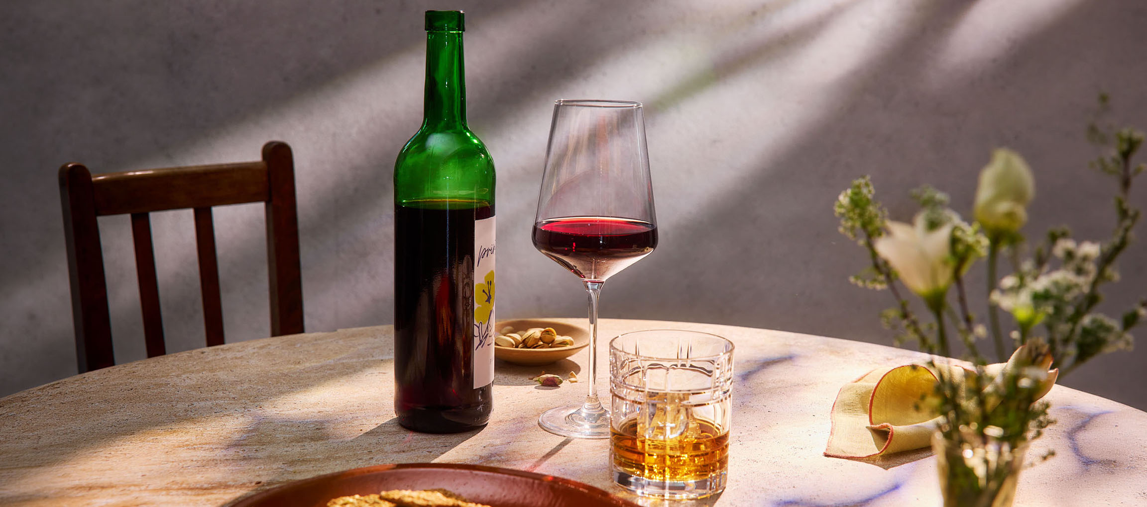 Fles rode wijn, wijnglas en whisky op tafel, met stoel en bloemen op de achtergrond.