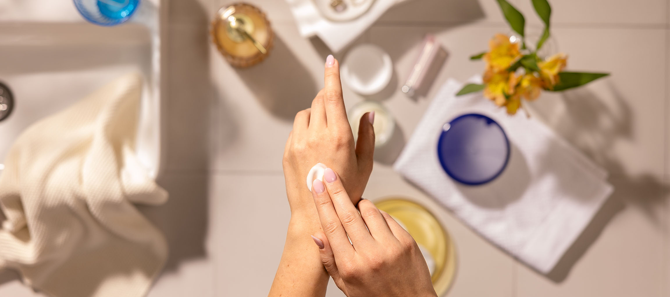 Vrouwenhanden smeren handcrème, omringd door cosmetica en handdoeken in een badkamer.