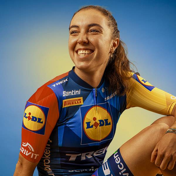 Vrolijke vrouw in een blauw, geel en rood fietsersshirt met logo's, lachend en kijkend naar rechts.